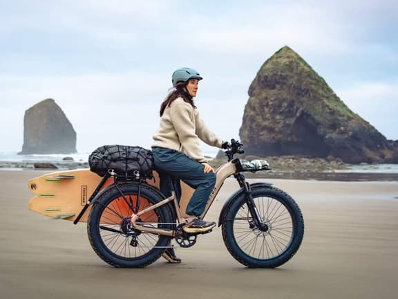 A rider on a sand-coloured Aventon Aventure fat tyre electric bike on a beach with a surfboard strapped to the rear rack