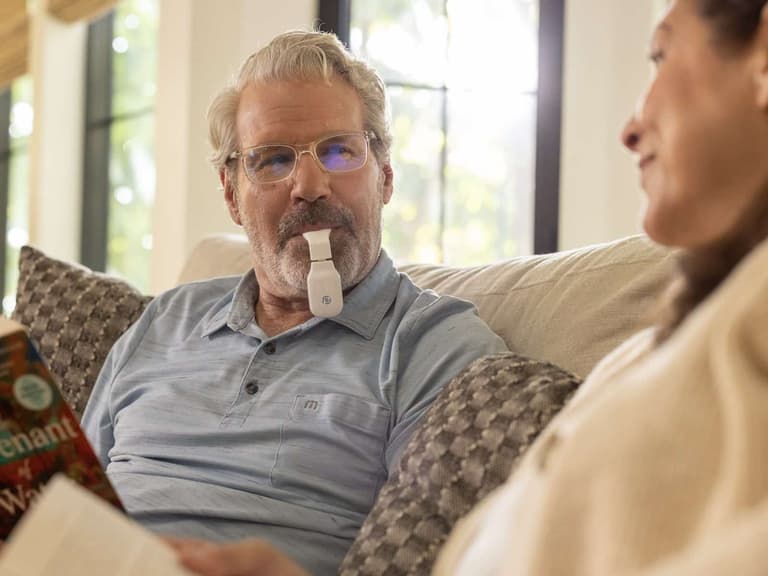 A man using the Spring Sleep eXciteOSA daytime therapy device while reading on the couch beside his partner.