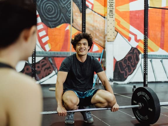 A smiling personal trainer crouched behind a loaded barbell talking to a client in a gym