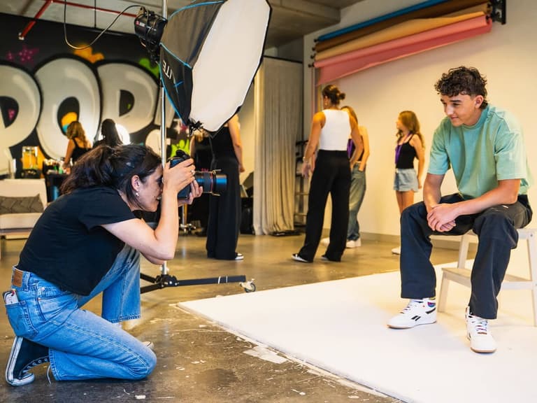 A POP Photography photographer crouches with a camera to shoot a teenage boy seated on a stool against a white studio backdrop, with a large softbox and a graffiti POP mural behind her