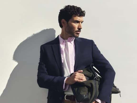 A man in a navy blazer, pink Sportscraft Oxford shirt and tan shorts holding a green weekender bag against a white wall