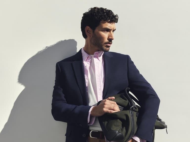 A man in a navy blazer, pink Sportscraft Oxford shirt and tan shorts holding a green weekender bag against a white wall
