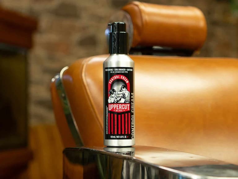 A silver aluminium bottle of Uppercut Deluxe Control Cream standing on the chrome arm of a tan leather barber chair against a stone wall.