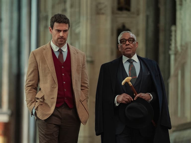 "Two well-dressed men walking together inside a grand, historic stone building with high ceilings and stained glass windows, suggesting a church or cathedral setting."