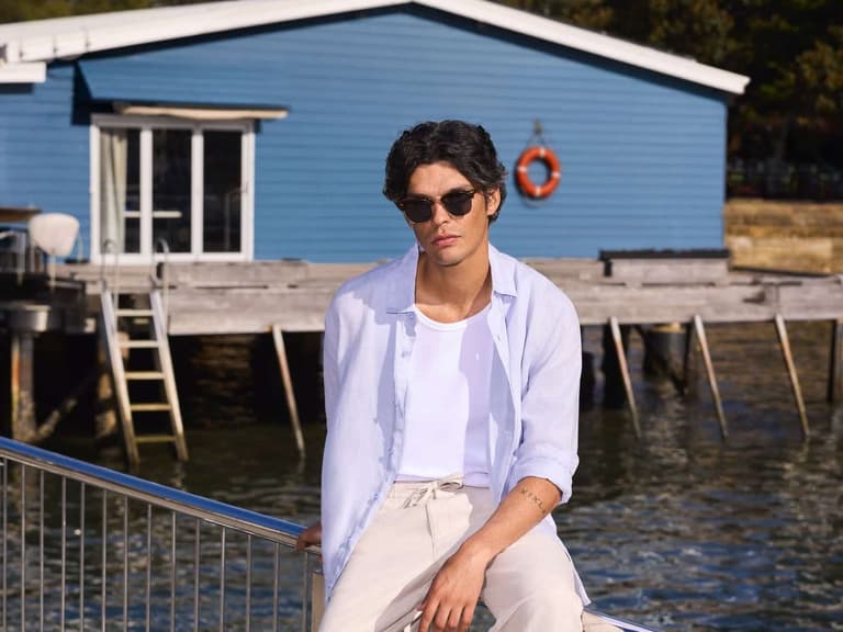 A man in sunglasses wearing a light blue long sleeve shirt over a white tee and cream pants, sitting on a waterfront railing in front of a blue boatshed
