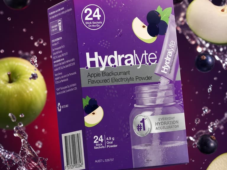 A purple 24 pack box of Hydralyte Apple Blackcurrant Stick Sachets surrounded by apple slices, blackcurrants and splashing water