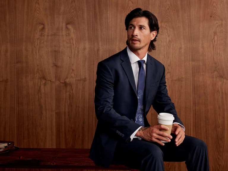A man in a navy suit and blue tie holding a coffee, from the Men's Wearhouse fall campaign