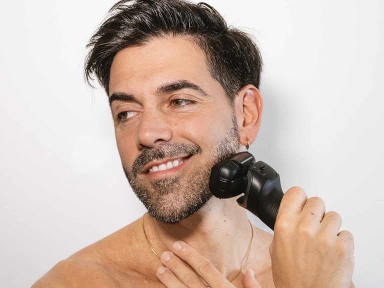 A man with stubble shaving his jawline with the black Panasonic Series 900 PRO ES-L580U electric shaver against a white wall