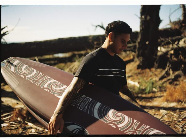 Kehu Butler carrying a SharpEye surfboard decorated with Tai Kehu artwork through coastal scrub