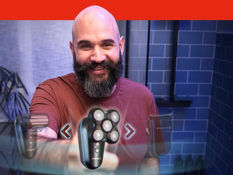 A bald man with a full dark beard holding the Remington Balder Head & Beard shaver towards the camera in a blue tiled bathroom