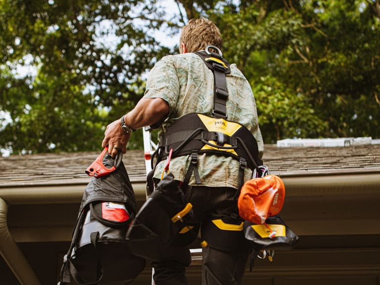 A roofer in a Petzl safety harness and Hawaiian shirt climbs a ladder onto a roof carrying gear bags
